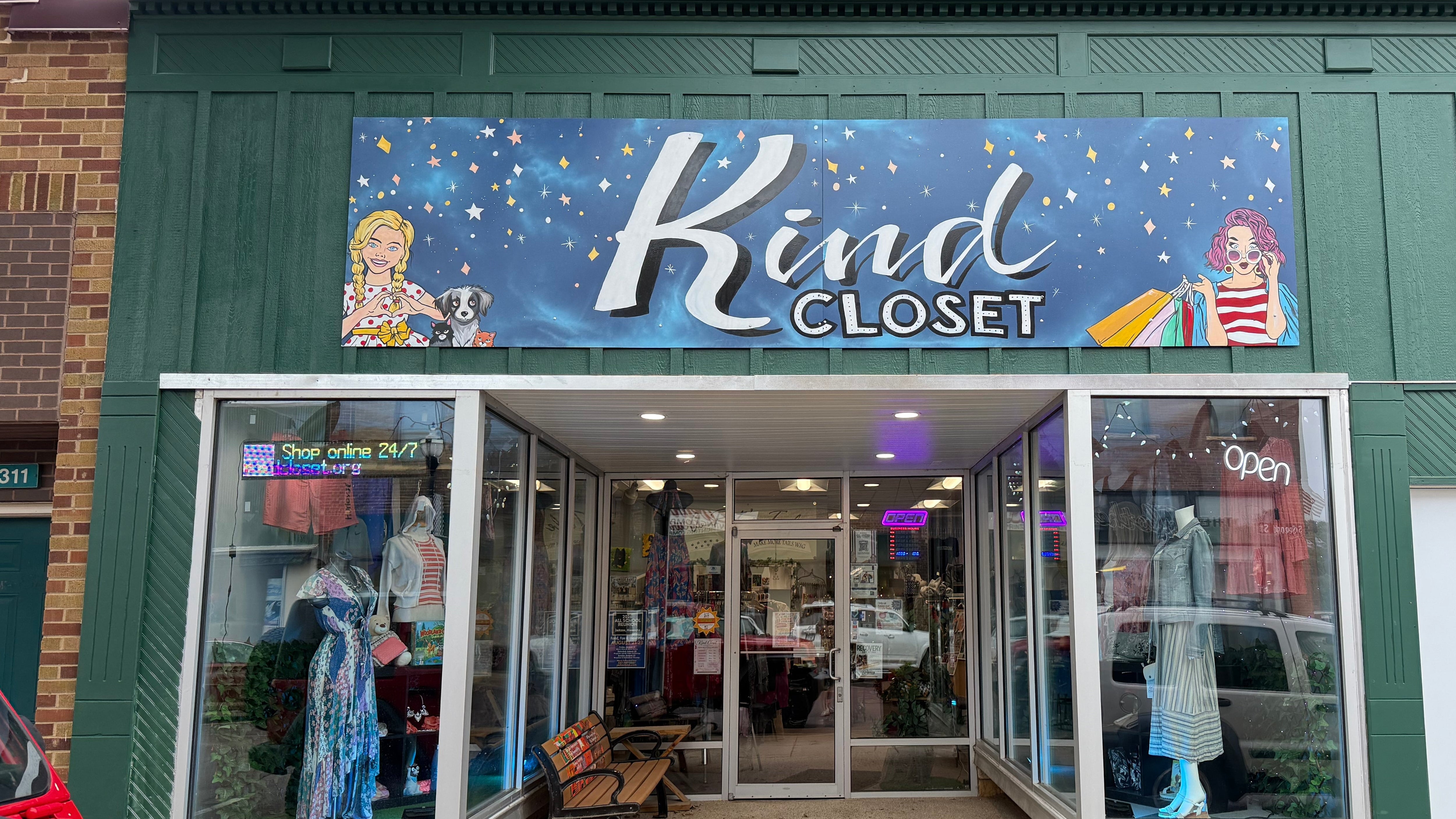 Front exterior of Kind Closet retail store with a large blue storefront sign, glass display windows, and a brick building facade. A red vehicle is partially visible in the foreground, and potted flowers sit near the entrance.